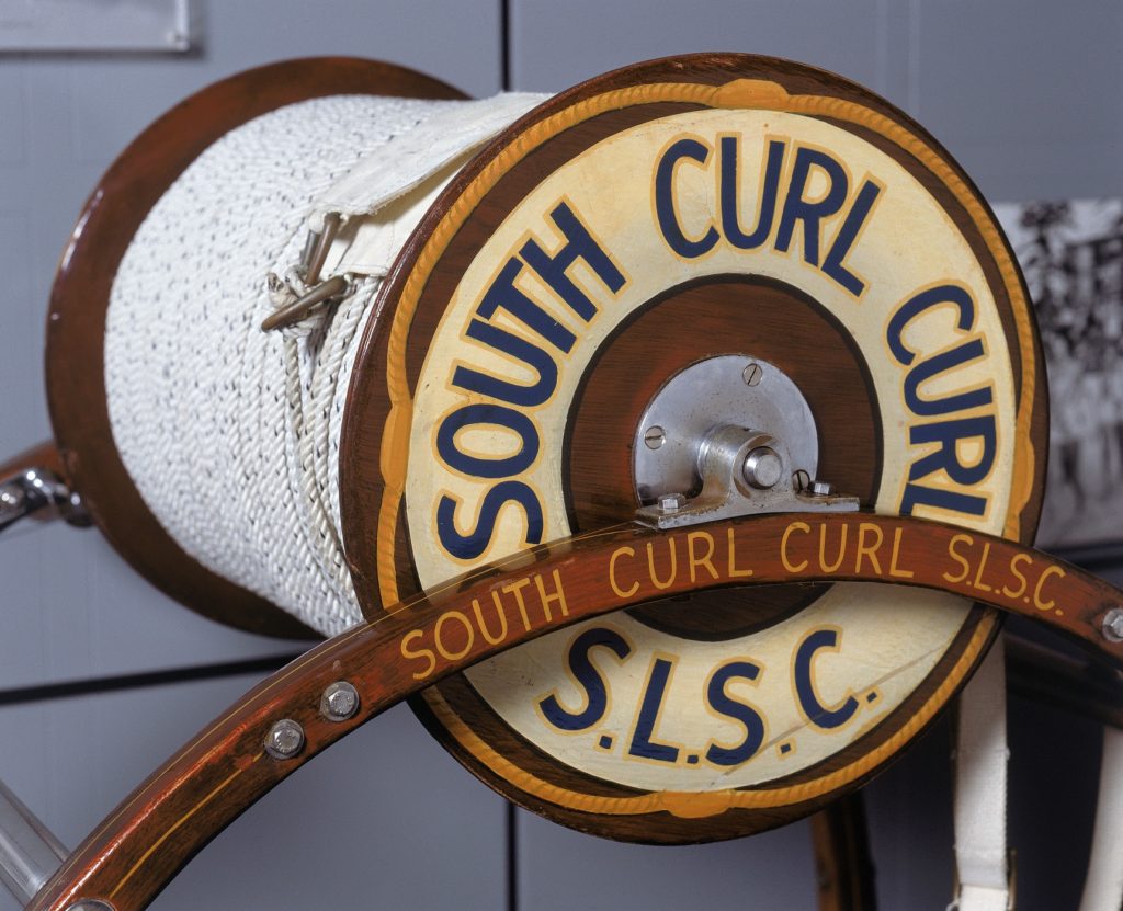 The reel comprises a large, polished wooden cylinder with cotton rope wound around it. The cylinder is set in a frame stand which could be carried to the water's edge. The name of the surf club is painted on the sides of the cylinder.